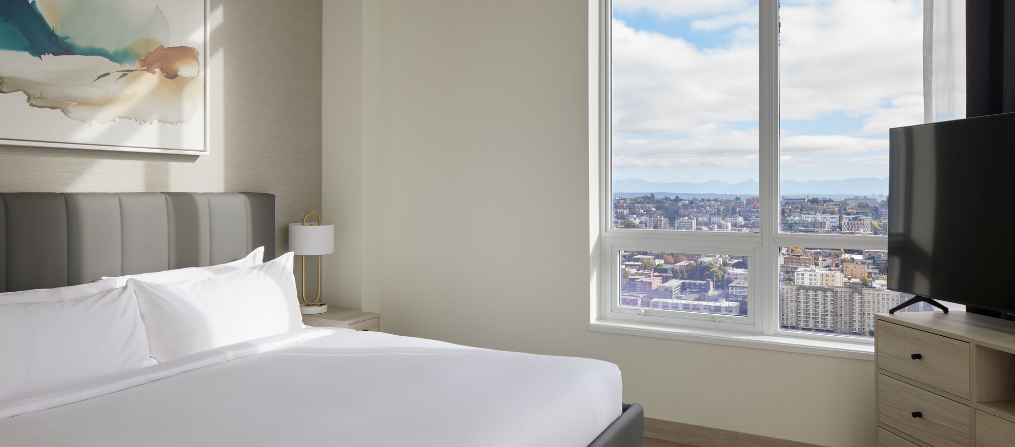 A modern, sunlit bedroom with a neatly made bed, light wood furniture, a large window offering a cityscape view, a flat-screen TV, and abstract wall art above the bed.
