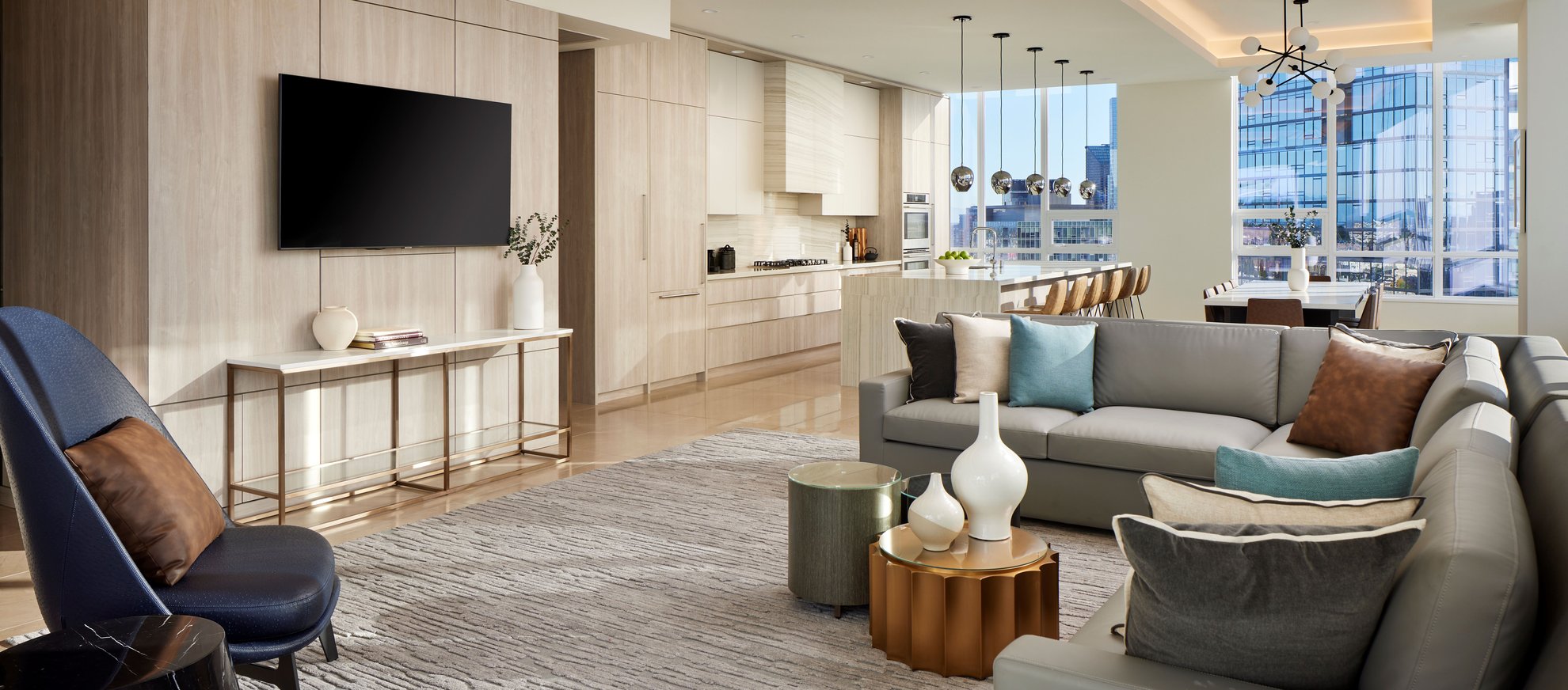 Modern open-concept living room and kitchen with light wood cabinets, a wall-mounted TV, neutral sofas with accent pillows, contemporary décor, bar stools at a kitchen island, and large windows with city views.