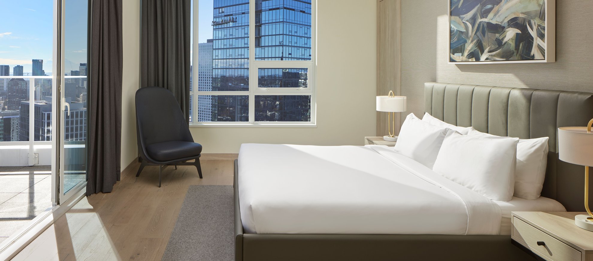 A modern hotel room with a large bed, white linens, bedside tables with lamps, a dark chair by the window, abstract wall art, wood floors, and a city view through large windows and a glass door.