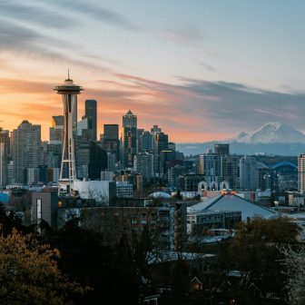 Seattle skyline at sunset, featuring the Space Needle, downtown skyscrapers, and Mount Rainier in the background under a partly cloudy sky. Trees and buildings are visible in the foreground.