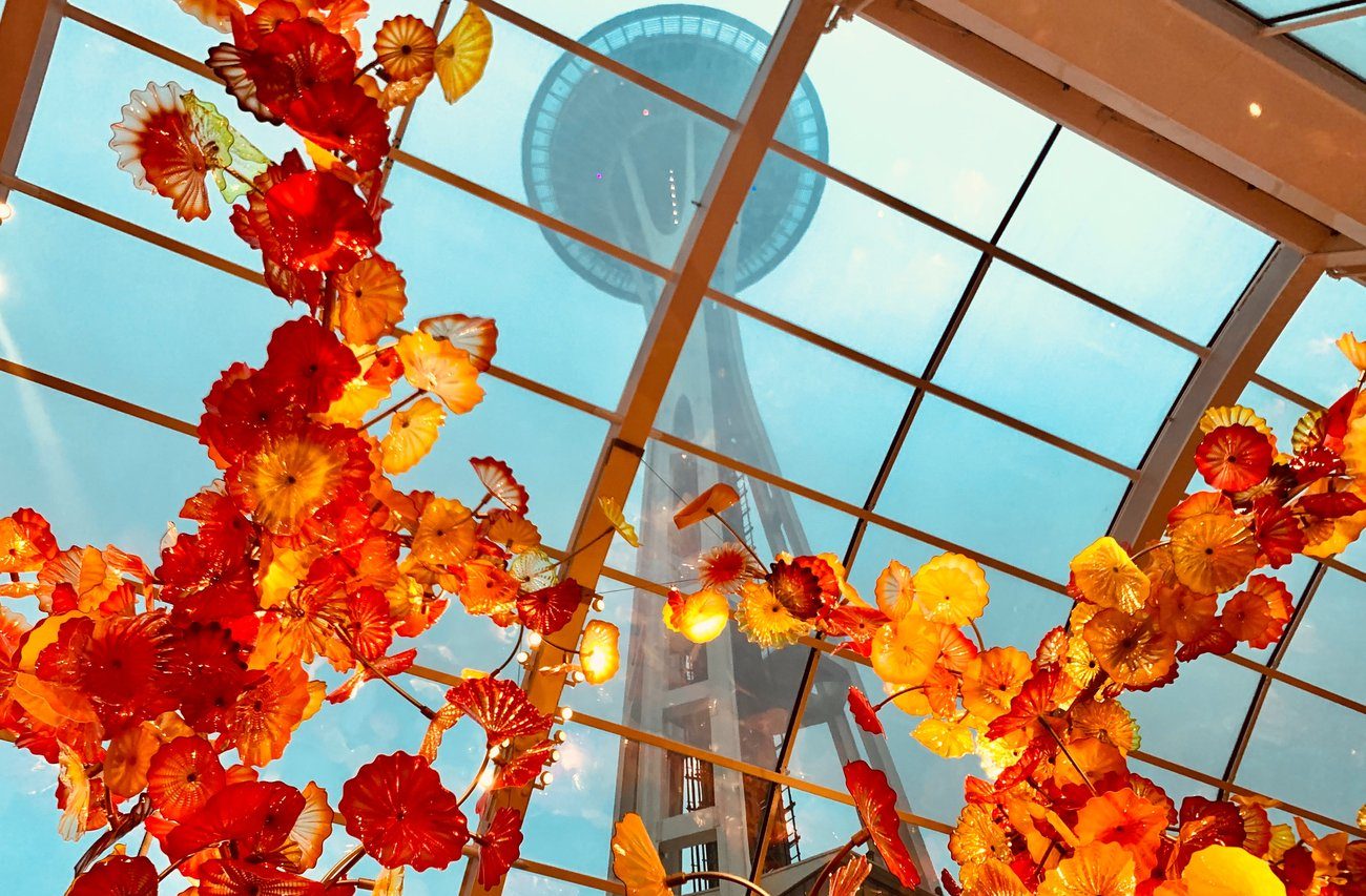 Bright orange and yellow glass sculptures hang from the ceiling inside a glass atrium, with the Space Needle visible through the windows above on a cloudy day.