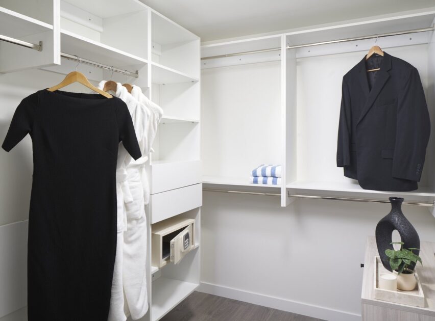 A tidy walk-in closet with white shelves, a black dress and white robe on hangers, a black suit jacket, folded towels, a black vase on a table, and a safe on a shelf.