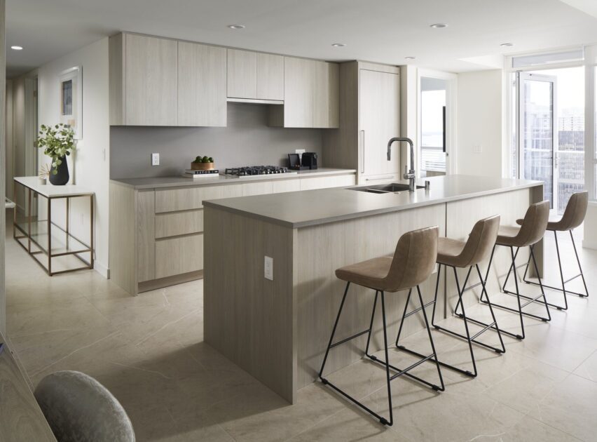 Modern kitchen with light wood cabinets, a large island with a built-in sink, and four tan barstools. There are built-in appliances, neutral countertops, and large windows letting in natural light. A hallway table with flowers is visible.