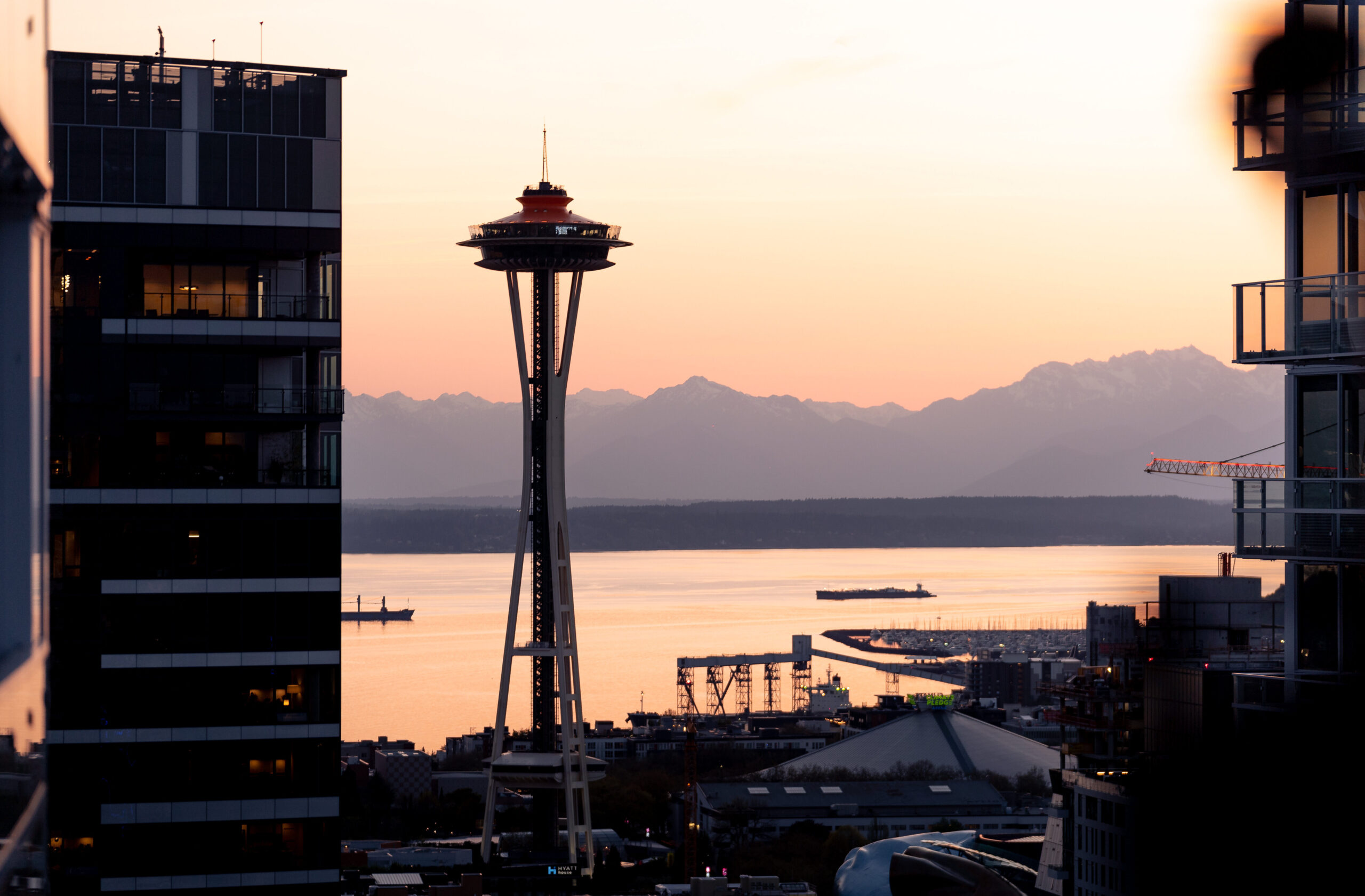 The Space Needle in Seattle stands against a pink and orange sunset sky, with mountains and water in the background, framed by tall buildings on either side.