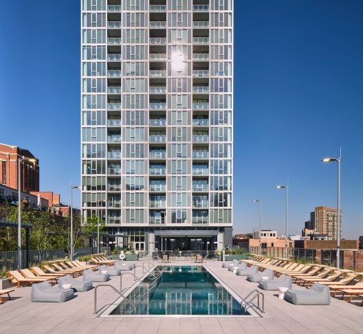 A modern high-rise apartment building with a rooftop pool surrounded by lounge chairs and city buildings under a clear blue sky.