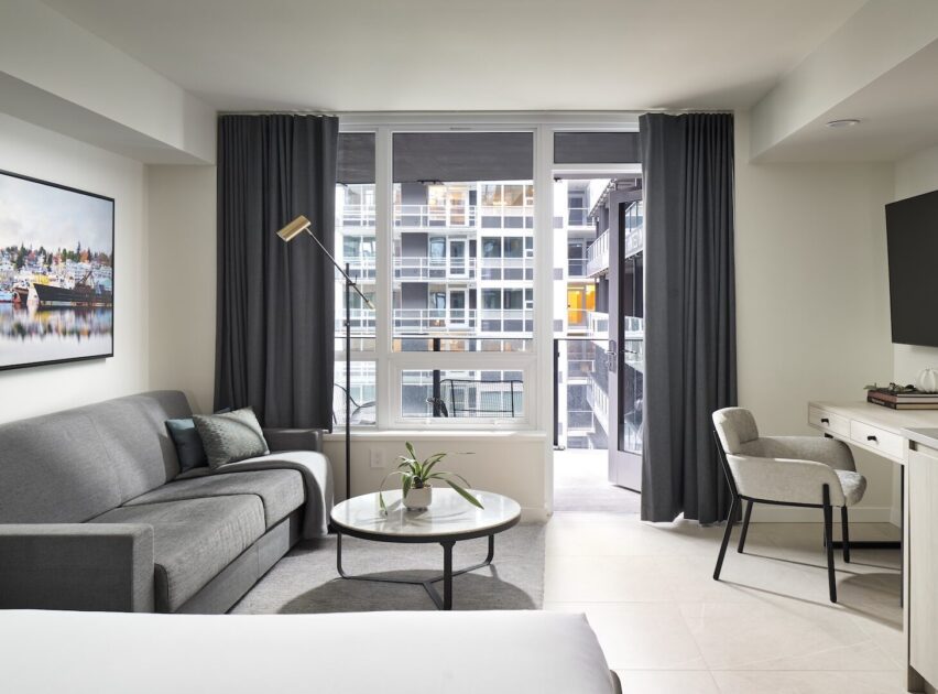 Modern hotel room with a gray sofa, round coffee table, floor lamp, desk with chair, large window with dark curtains, and a view of an urban apartment building outside. A potted plant and wall art add decorative touches.