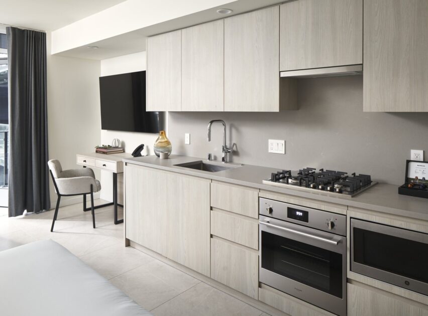 A modern kitchen with light wood cabinets, built-in oven, gas stove, microwave, sink, and a gray countertop. In the background is a small desk with a chair and a TV mounted on the wall.