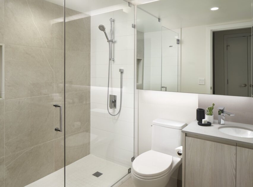 Modern bathroom with a glass-enclosed shower, beige tile walls, white toilet, light wood vanity with a sink, and minimal decor including a soap dispenser and cup. The space is bright and clean.