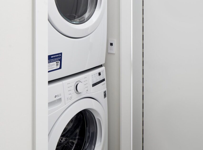 A white stacked washer and dryer unit is installed in a small laundry closet with white walls and a partially open sliding door. The control panels and a wall switch are visible.
