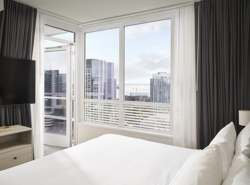 A modern hotel room with a neatly made bed, large windows with sheer white and dark curtains, a flat-screen TV, and a cityscape view with tall buildings outside.