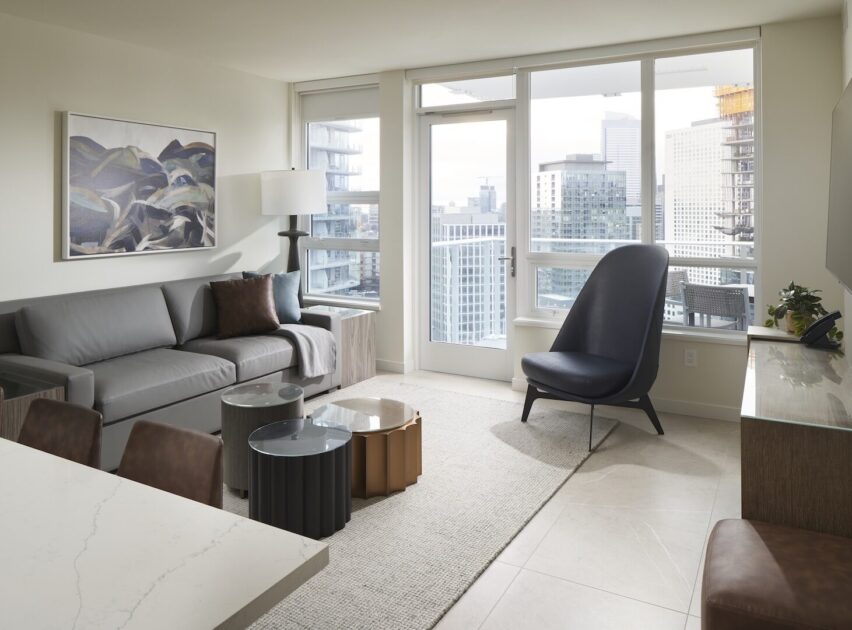 Modern living room with a gray sofa, unique coffee tables, a dark accent chair, TV on a console, large windows, and a city skyline view. Neutral tones and contemporary decor create a bright, stylish space.