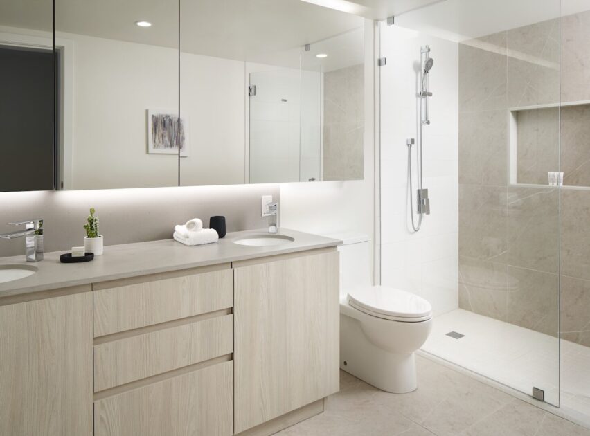 Modern bathroom with light wood vanity, double sinks, large mirror, white toilet, and a spacious glass-enclosed shower with beige tiles. Towels and a small plant sit on the counter; recessed lighting brightens the space.
