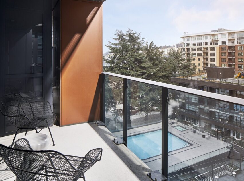 A modern balcony with black wire chairs overlooks an outdoor swimming pool and surrounding apartment buildings, with tall trees and a partly cloudy sky in the background.