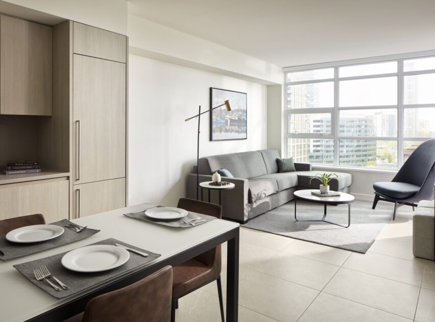 Modern apartment interior with a dining table set for two in the foreground, a gray sofa, round coffee table, and large windows allowing natural light into the living area with city buildings visible outside.