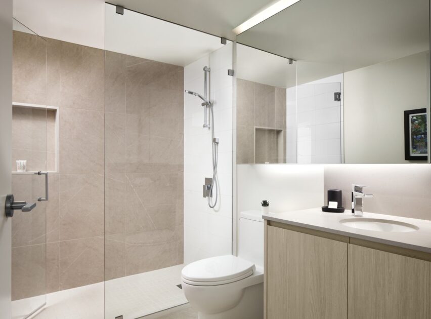 Modern bathroom with a glass-enclosed shower, beige and white tiled walls, a wall-mounted showerhead, a white toilet, a light wood vanity with a sink, and a large mirror above the counter.
