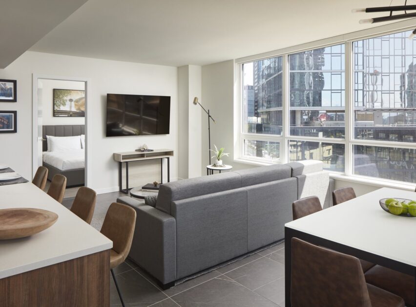 Modern apartment interior with gray sofa, wall-mounted TV, dining area, and kitchen island; large windows provide city views and natural light; a bedroom is visible through an open door.