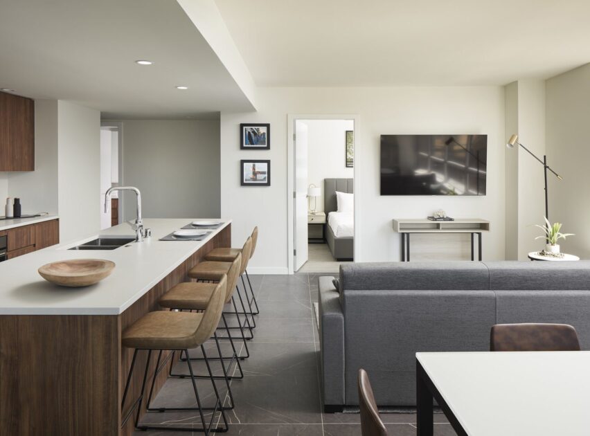 Modern open-plan living space with a kitchen island and barstools, a gray sofa facing a wall-mounted TV, a dining table, and a view into a bright bedroom through an open door. Neutral tones and minimalist decor.