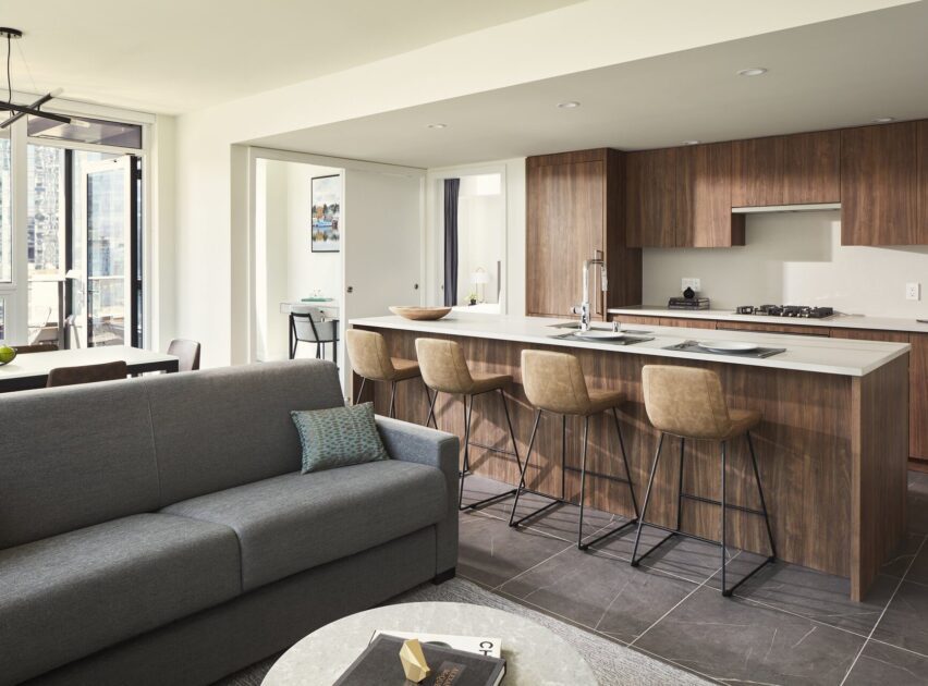 Modern open-concept kitchen and living area with a gray sofa, round coffee table, wooden kitchen island with four barstools, and built-in appliances. Large windows provide natural light.