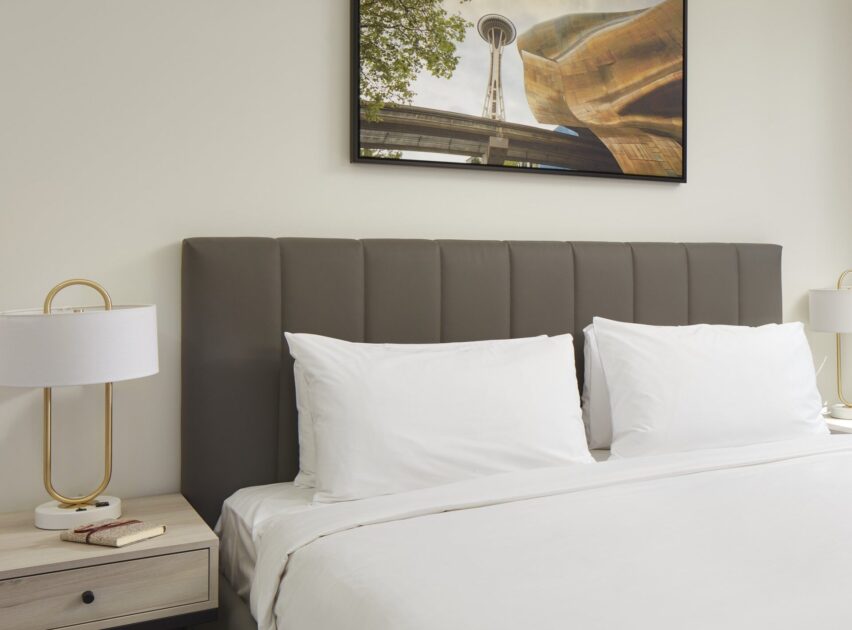 A neatly made bed with white linens and two pillows stands against a beige wall. Above the bed hangs a framed photo of Seattle’s Space Needle. Two bedside tables each have a modern lamp with a gold base.