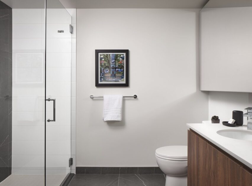 Modern bathroom with a glass shower, white walls, a towel hanging on a rail under a framed picture, a toilet, and a countertop with a sink and toiletries. Dark gray floor tiles and wood cabinet.