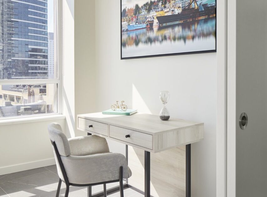 A modern, minimal desk with a cushioned chair sits by a bright window. Items on the desk include an hourglass, a decorative object, and a framed photo of boats hangs on the wall above.
