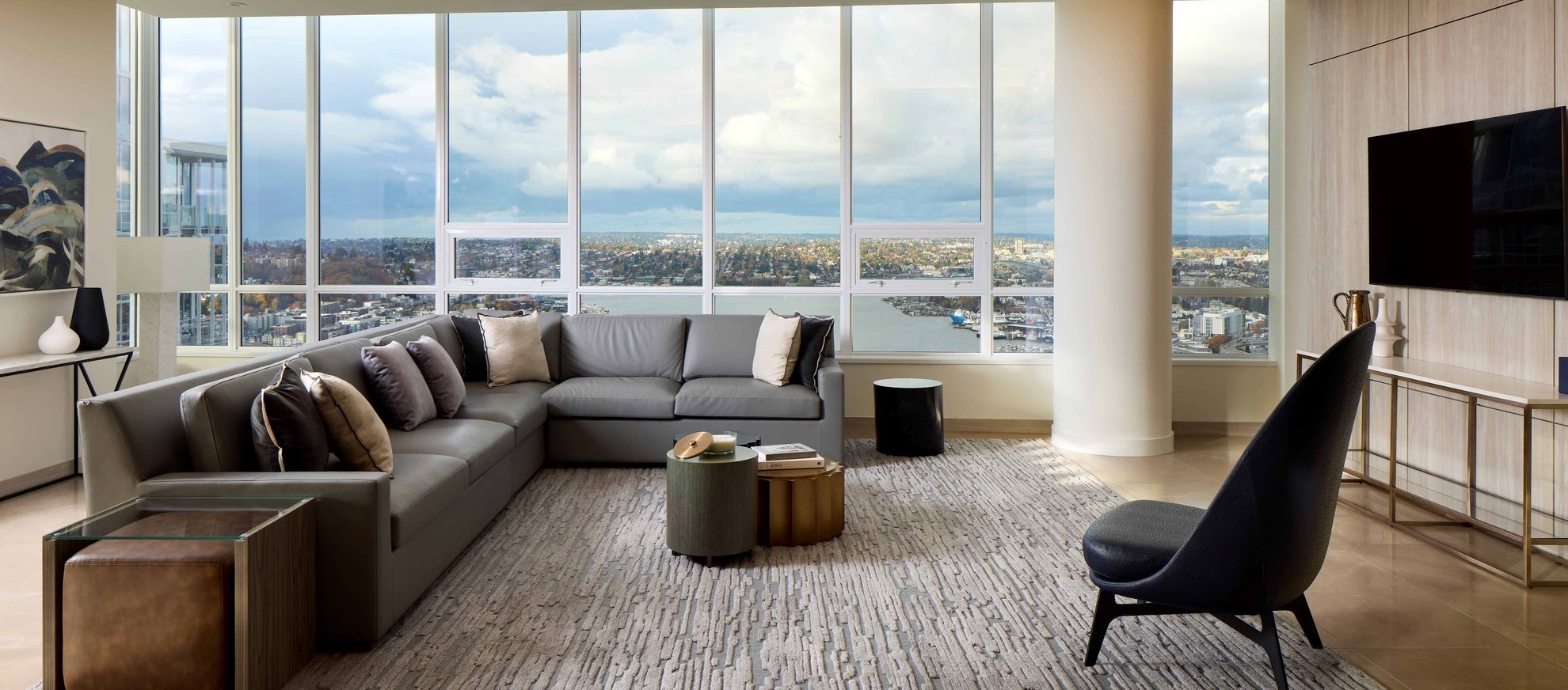 Modern living room with large windows showing a city and water view, gray sectional sofa with beige pillows, accent chairs, coffee tables, and neutral decor on a textured rug. Natural light fills the space.