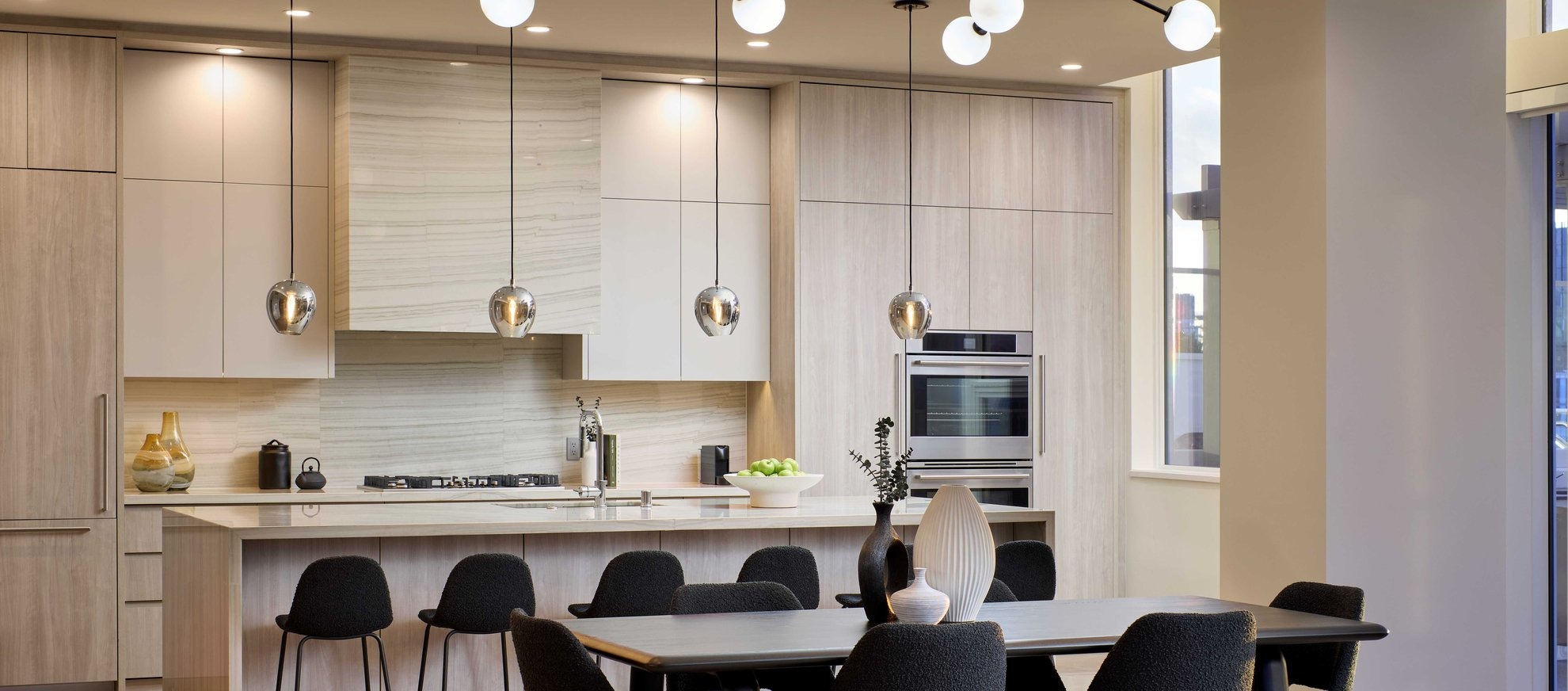 Modern kitchen with light wood cabinets, a marble backsplash, and built-in oven. Pendant lights hang over a kitchen island with bar stools. A dining table with black chairs and decorative vases is in the foreground.