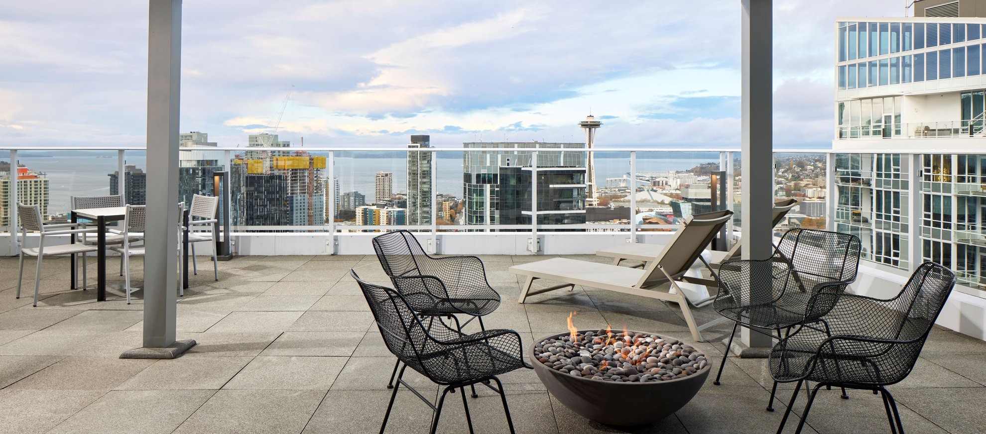 A rooftop patio with metal chairs around a fire pit, lounge chairs, and tables overlooks a city skyline, including the Space Needle and waterfront, under a partly cloudy sky.
