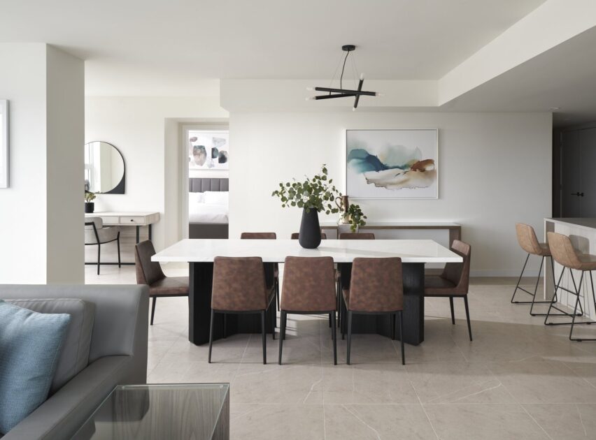 Modern dining room with a white table, brown chairs, a black vase with greenery, abstract artwork on the wall, minimalist decor, bar stools at a counter, and neutral tones throughout the space.