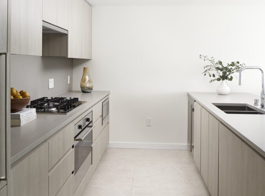Modern kitchen with light wood cabinets, a built-in oven, gas stove, and gray countertops. There are vases with greenery and a bowl of lemons on the counter, with a stainless steel sink and minimalist decor.