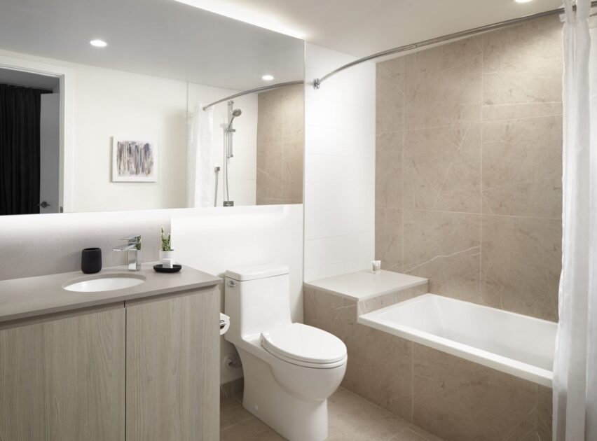 Modern bathroom with light wood vanity, white sink, and toilet. Bathtub with beige tile walls, curved shower rod, and white shower curtain. Minimalist decor, including a small plant and wall art, under recessed lighting.