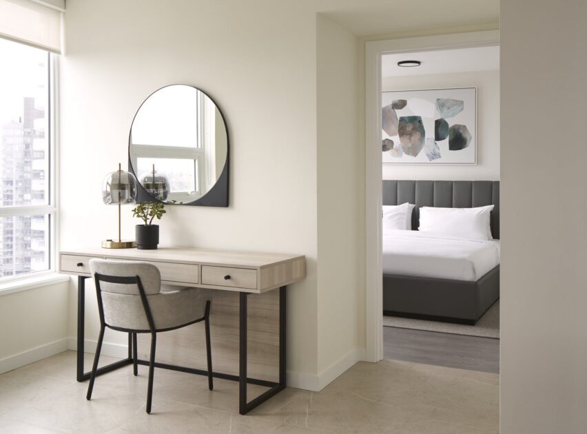 A modern, minimal home office features a light wood desk with a round mirror, black lamp, and potted plant. A gray upholstered chair sits at the desk. Through an open doorway, a tidy bedroom with a dark bed and abstract art is visible.