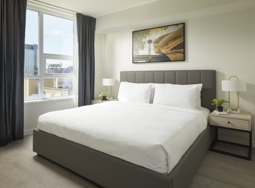 A modern, bright bedroom with a large bed, white bedding, two nightstands with lamps and plants, a large window with city views, dark curtains, and framed art above the headboard.