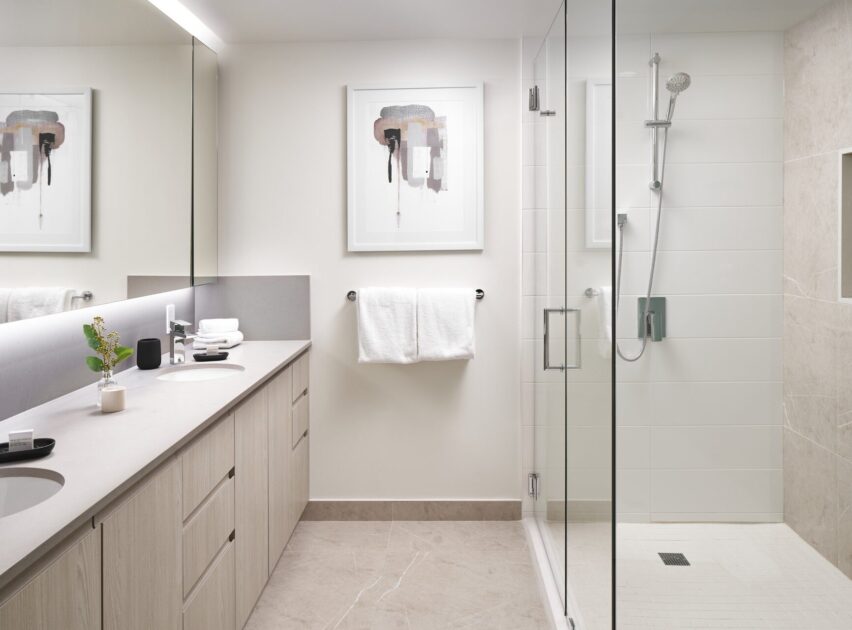 Modern bathroom with light wood cabinets, double sinks, a large mirror, a glass-enclosed shower, white towels, beige tile floor, and abstract art on the wall above the towel rack.