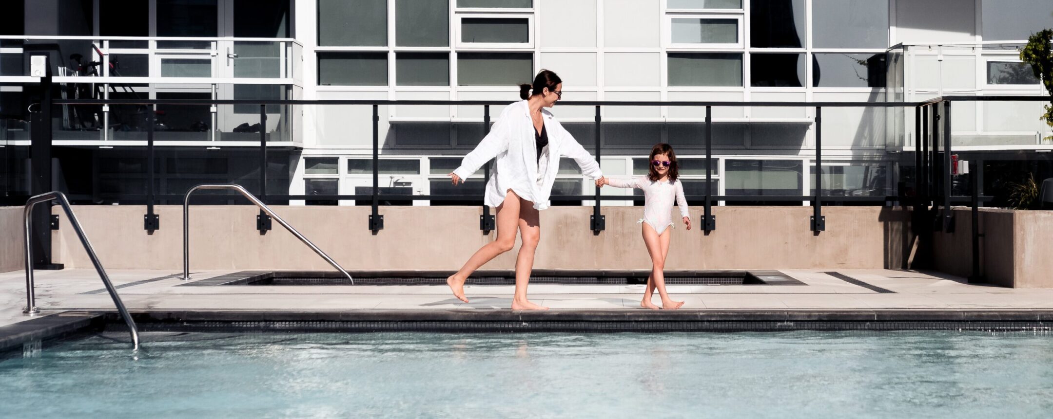 A woman and a young girl, both in swimwear and cover-ups, walk hand-in-hand beside a pool with a modern apartment building in the background. The scene is bright and sunny.