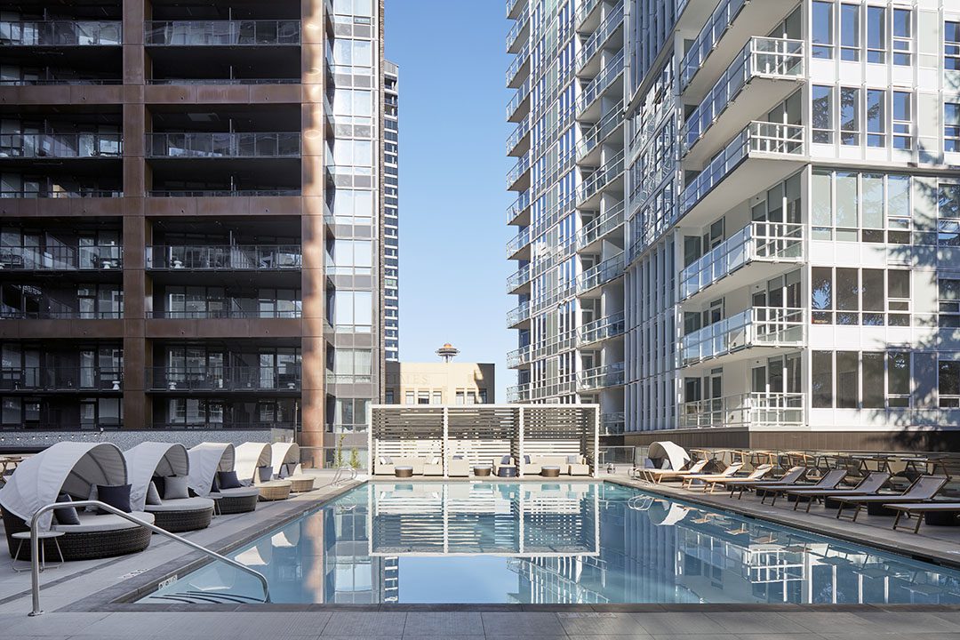 Modern outdoor swimming pool surrounded by lounge chairs and cabanas, situated between two tall apartment buildings with glass balconies, under a clear blue sky. The Space Needle is visible in the background.