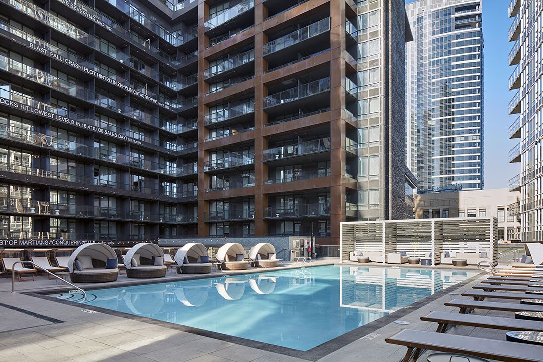 A modern outdoor pool surrounded by lounge chairs and round cabanas, set between tall glass and steel apartment buildings in an urban high-rise complex on a clear day.