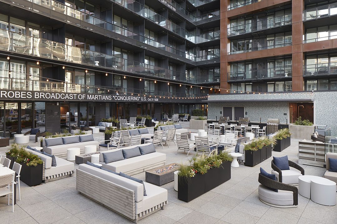 Modern outdoor patio with gray and white seating, planters, and string lights, surrounded by a multi-story building with balconies. A marquee sign displays a message in the background.