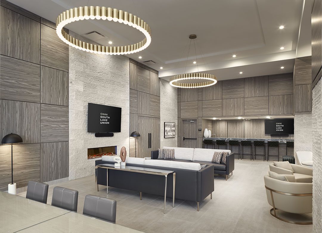 Modern lounge with wood-paneled walls, large circular chandeliers, two sofas, wall-mounted TVs, a fireplace, high kitchen cabinets, bar stools, and a dining table in the foreground. Neutral, elegant color palette.