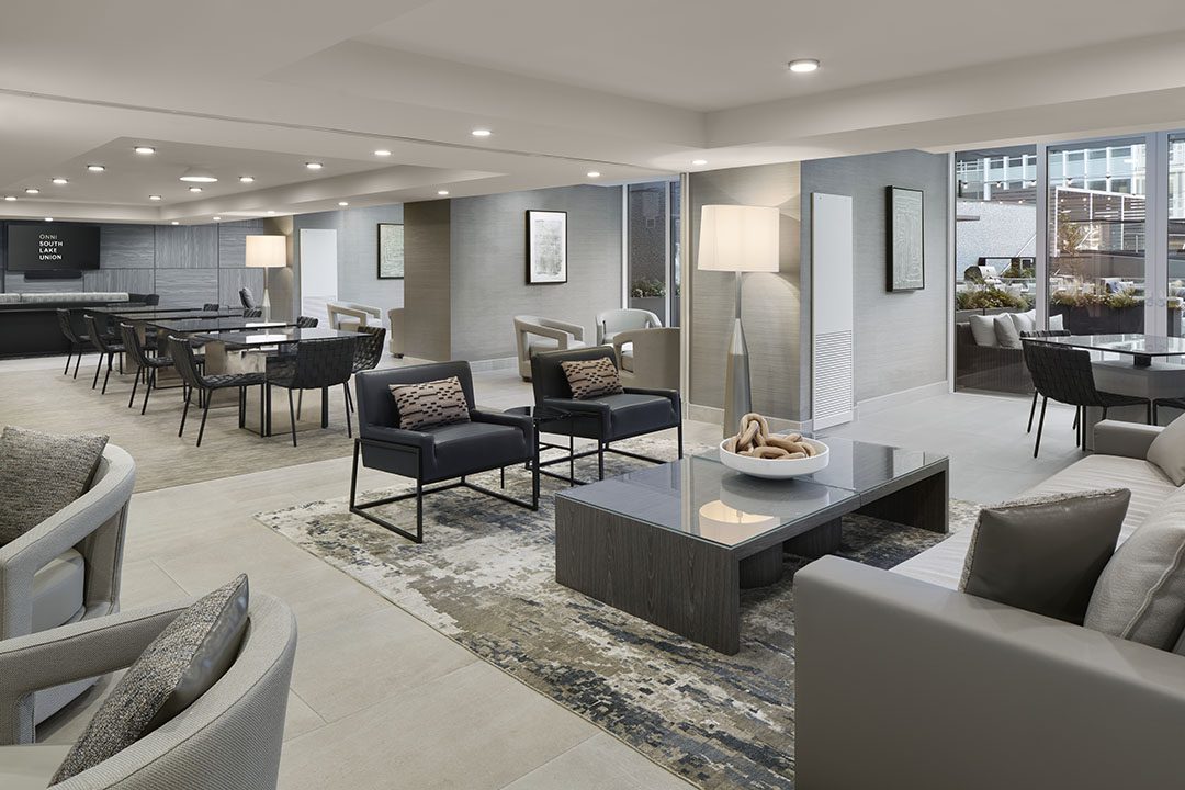 Modern lounge with neutral colors, featuring armchairs, sofas, a large rug, coffee tables, dining tables, and floor lamps. Large windows reveal an outdoor patio and city buildings in the background.