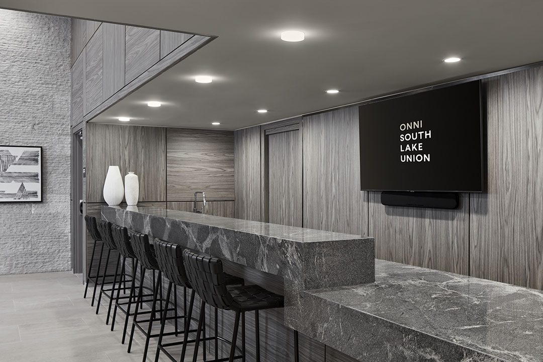 A modern, sleek lounge with a long gray marble counter, high black chairs, minimalist decor, and a wall-mounted TV displaying ONNI SOUTH LAKE UNION. Neutral tones and recessed lighting create a sophisticated atmosphere.