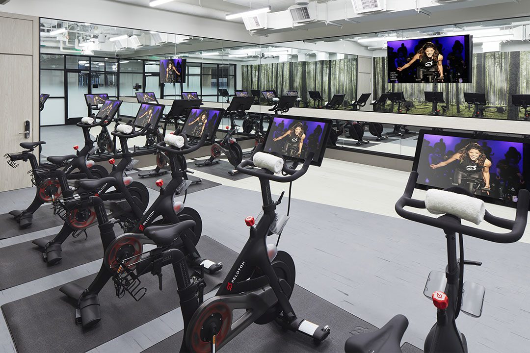 A modern indoor cycling studio with several stationary bikes, each equipped with a screen. A large mirror and TV display an instructor leading a class. Bright, clean space with towels on each bike.