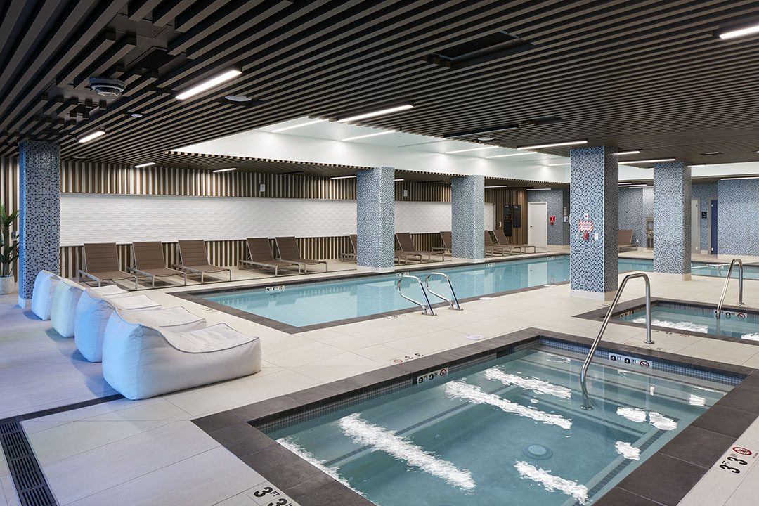 Modern indoor pool area with a small hot tub in the foreground, a larger swimming pool in the background, lounge chairs along the side, and striped wall and ceiling designs. The space is well-lit and clean.