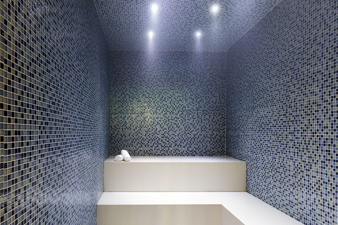 A modern steam room with blue mosaic tile walls and ceiling, built-in white seating, and three rolled white towels on the bench under soft ceiling lights.