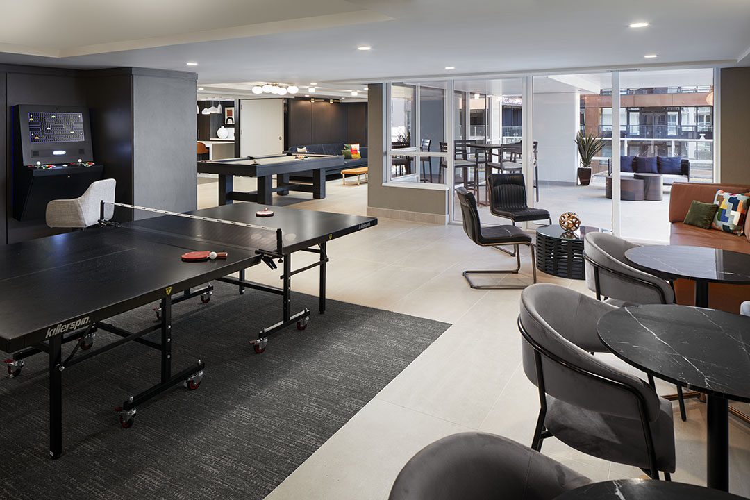 A modern game room with a ping pong table, pool table, arcade machine, lounge seating, and large windows overlooking a patio area with plants and outdoor furniture.