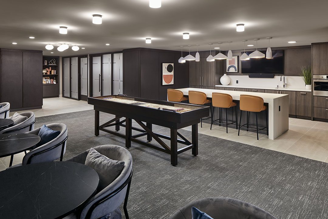 Modern communal lounge featuring a shuffleboard table, bar-style seating with orange chairs at a kitchen island, round tables with cushioned chairs, dark cabinetry, and contemporary lighting.