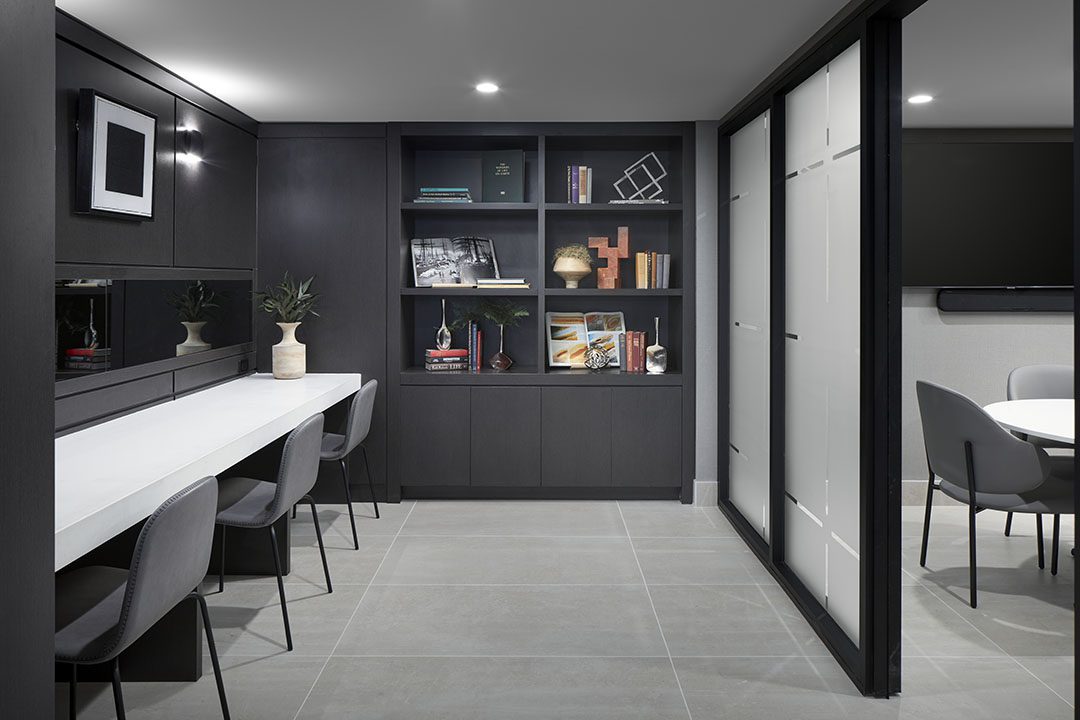 Modern office space with dark built-in shelves holding books and decor, a long white desk with gray chairs, and a meeting area with a round table and chairs behind a glass partition. The room is well-lit and minimalist.
