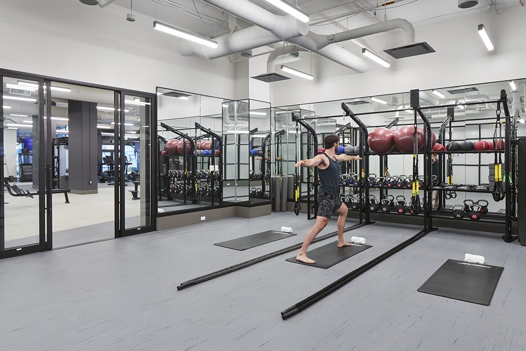 A man practices yoga on a mat in a modern gym with large mirrors, fitness equipment, medicine balls, and dumbbells. The space is clean, bright, and features glass walls and an organized layout.