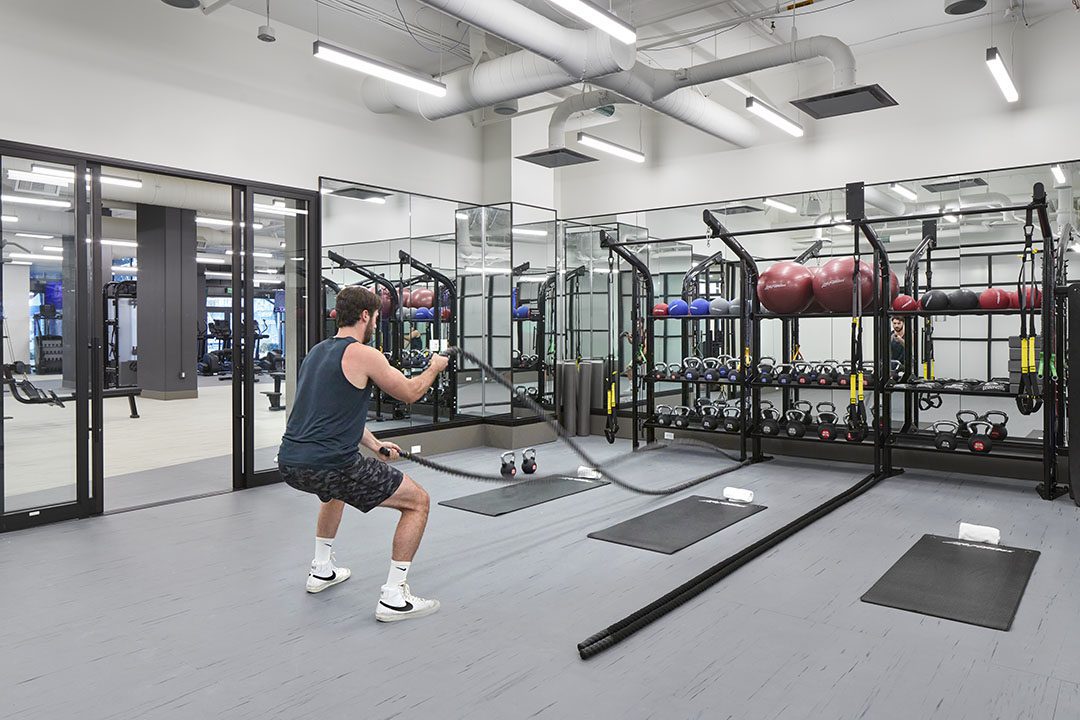 A man in athletic wear exercises with battle ropes in a modern gym. The gym has large mirrors, racks with medicine balls, kettlebells, and various workout equipment neatly arranged.