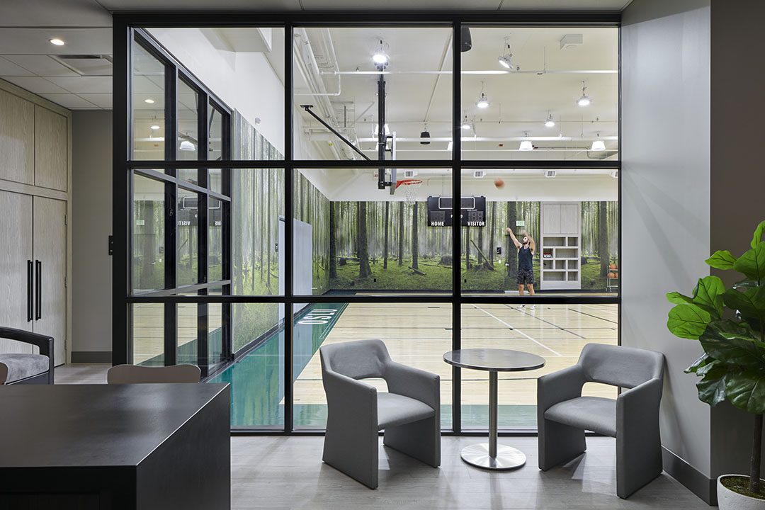 A modern indoor basketball court with a forest mural on the walls, viewed through large glass windows from a lounge area with two gray chairs and a small round table. One person is shooting a basketball.